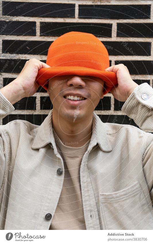 Positiver Mann setzt orangefarbenen Hut auf den Kopf positiv angezogen Augen abdecken Glück Kopfbedeckung cool ziehen Backsteinwand heiter Hipster Mode Spaß