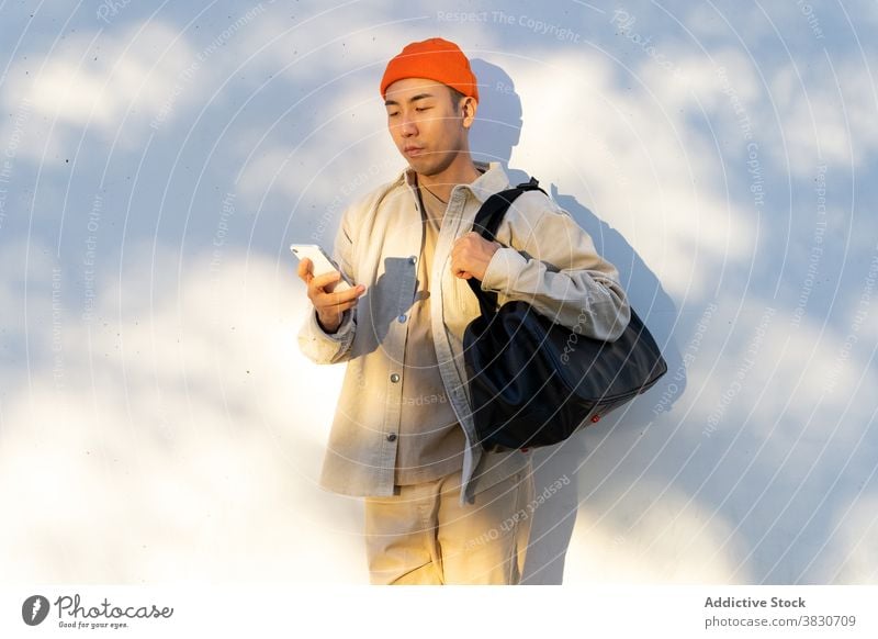 Asiatischer Mann mit Reisetasche mit Smartphone gegen weiße Wand benutzend Konzentration Browsen Funktelefon Fokus Surfen Apparatur modern jung lässig Mobile