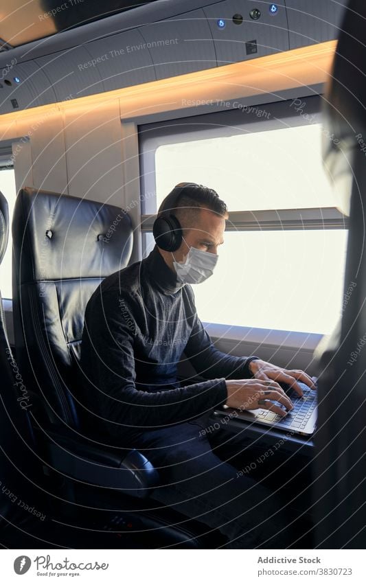 Mann mit Maske benutzt Laptop im Zug benutzend freiberuflich Projekt Mundschutz Pandemie zuhören Musik männlich jung Kopfhörer reisen Seuche COVID19 Bund 19
