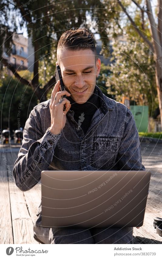 Fröhlicher junger Mann arbeitet am Laptop und spricht am Telefon benutzend sprechend Tippen heiter Internet freiberuflich Browsen Beruf männlich Arbeit