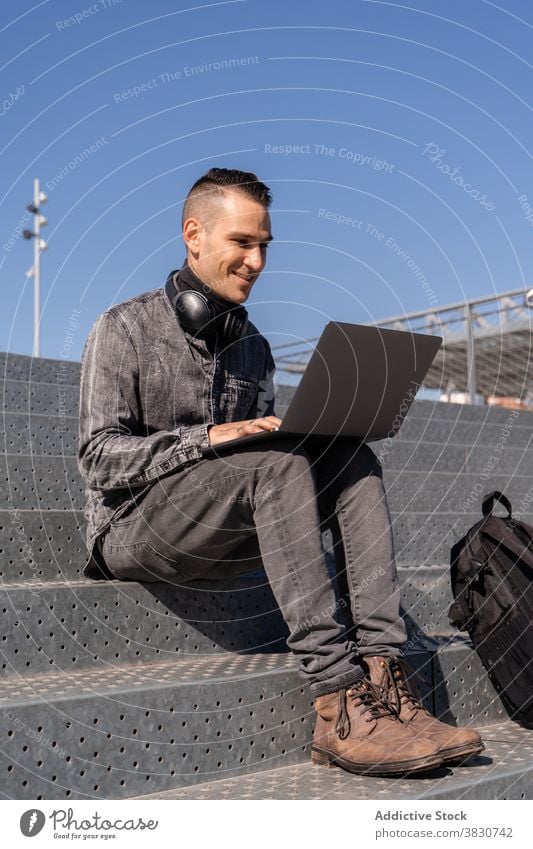 Fröhlicher junger Mann arbeitet am Laptop mit Kopfhörern benutzend Tippen heiter Internet freiberuflich Browsen Treppe Beruf männlich Arbeit abgelegen Gerät