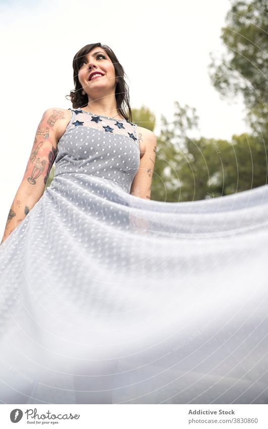 Fröhliche Frau in Kleid im Park in der Tageszeit fliegen informell heiter Tattoo Mode traumhaft Stil schlanke Zahnfarbenes Lächeln Inhalt froh Glamour