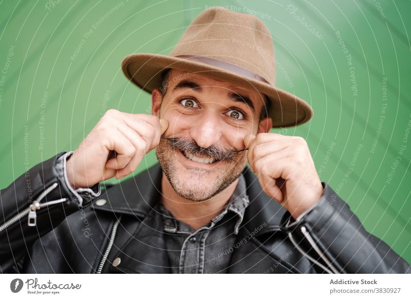 Fröhlicher Mann mit Schnurrbart im Hut und Grimasse Locken Gesicht machen Spaß heiter Lederjacke männlich froh Lächeln so tun, als ob Metrosexualität Optimist