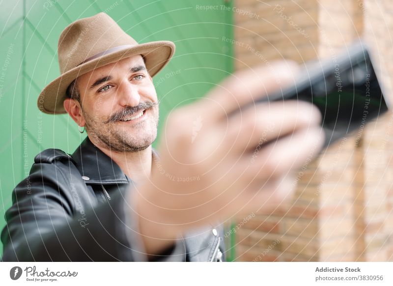 Erwachsener Mann mit Schnurrbart nimmt Selfie heiter Hipster trendy Vollbart Kavalier Hut Metrosexualität Lederjacke Hof Inhalt Typ Smartphone Hobby