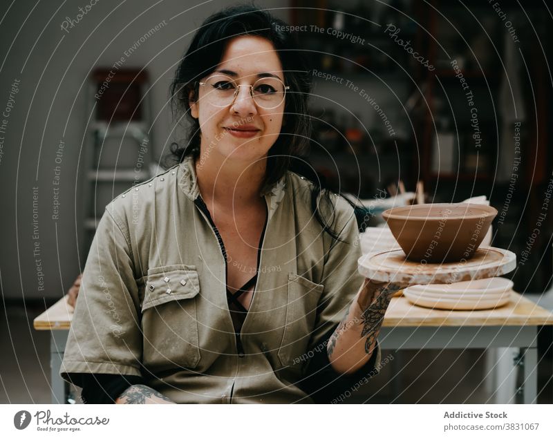 Lächelnde Keramikerin in der Werkstatt sitzend Handwerkerin Kunstgewerbler Ton Töpferwaren Sammlung Steingut Geschirr Teller Talent Frau hölzern Regal