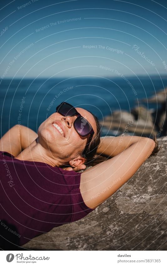 Liegende Frau auf einem Felsen, an der Küstenlinie mit Sonnenbrille und lila T-Shirt, im Hintergrund das Meer. Lächeln Wellen Wasser liegend Waffen Lügen