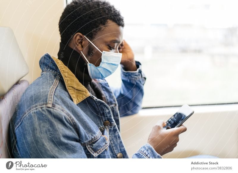 Reisender schwarzer Mann benutzt Handy im Zug Passagier Smartphone neue Normale reisen Mundschutz Coronavirus Eisenbahn männlich ethnisch Afroamerikaner Tourist