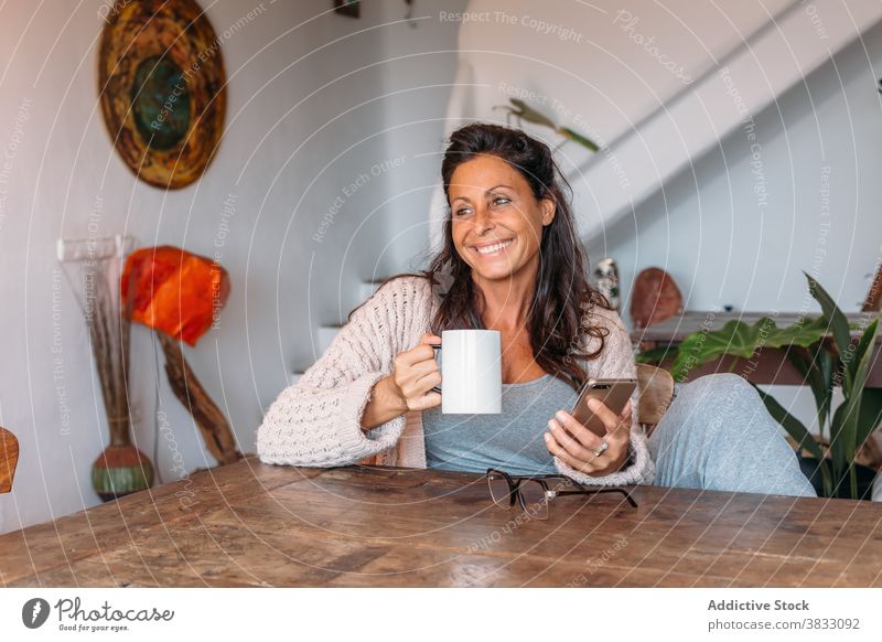 Glückliche Frau mit Tasse Kaffee und Smartphone sitzt am Tisch zu Hause heiter ruhen trinken benutzend Erwachsener ethnisch Mobile Telefon Gerät Apparatur