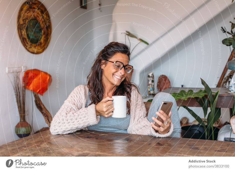 Positive ethnische Frau mit Tasse Kaffee beim Surfen auf dem Smartphone zu Hause Glück heiter ruhen trinken benutzend gemütlich Komfort Erwachsener Mobile
