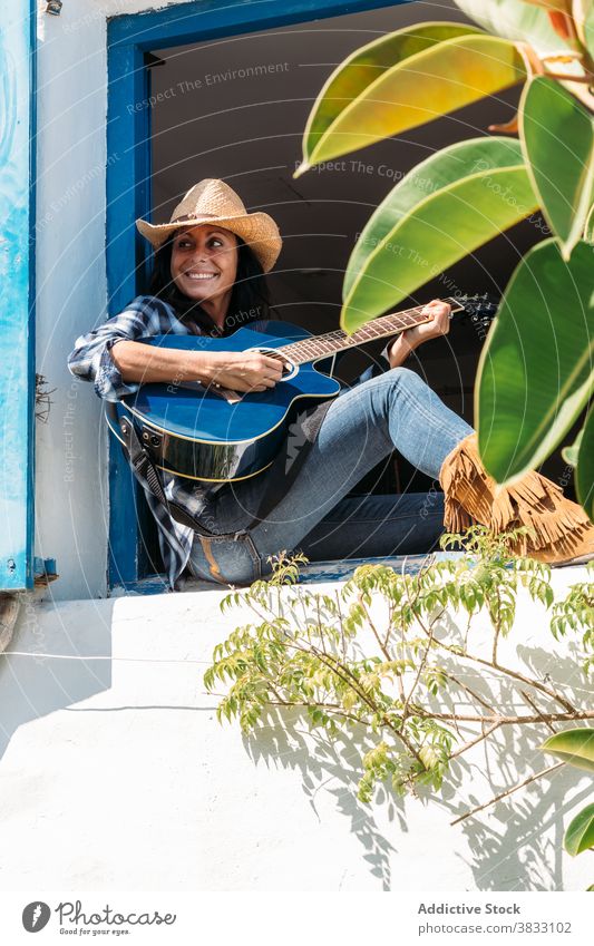 Glückliche Frau mit Cowboyhut und Stiefeln spielt Gitarre auf der Fensterbank Land ländlich spielen Kälte ruhen Stil heiter ethnisch Fenstersims Landschaft