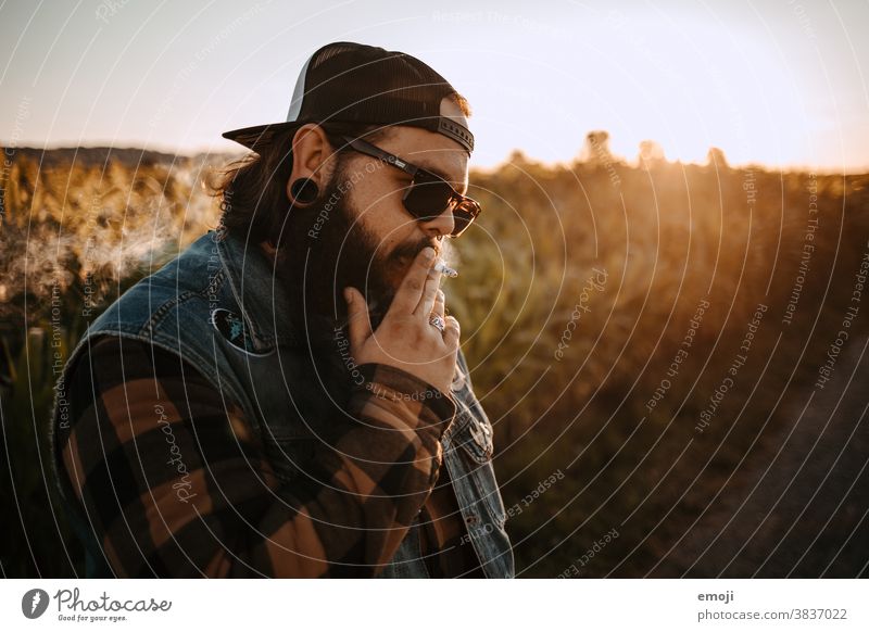 junger Mann mit Bart, Sonnenbrille und Cap raucht jugendlich sonnenuntergang Draussen feld trendy Hipster Rauchen Zigarette Kopfbedeckung ungesund Lifestyle