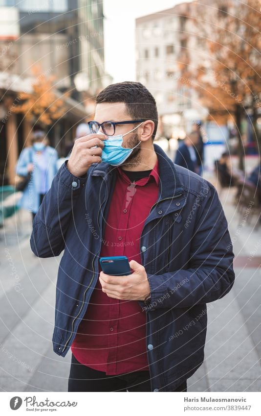 Mann repariert die Gesichtsmaske, während er in der Stadt spazieren geht und ein Smartphone in der Hand hält Erwachsener Großstadt Mitteilung Coronavirus