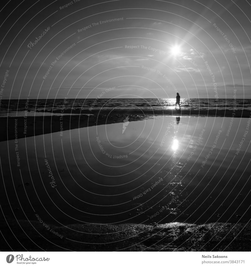 einsame Frau, die am Ostseestrand bei hochsommerlichem Sonnenuntergang über dem Wasser spazieren geht. Schwarz und weiß. täglich Leben anonym Rücken Strand