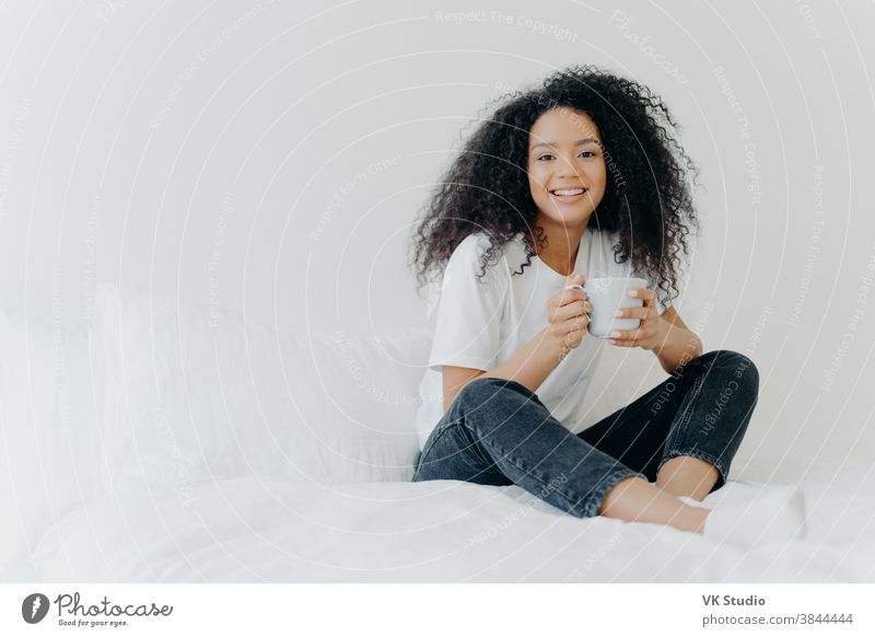 Junge hübsche afroamerikanische Frau ruht zu Hause im Bett, trinkt morgens heißen Tee, genießt häusliche Atmosphäre, leckeres Getränk, während sie sich im Schlafzimmer entspannt. Menschen, Freizeit-, Ruhe- und Lifestyle-Konzept