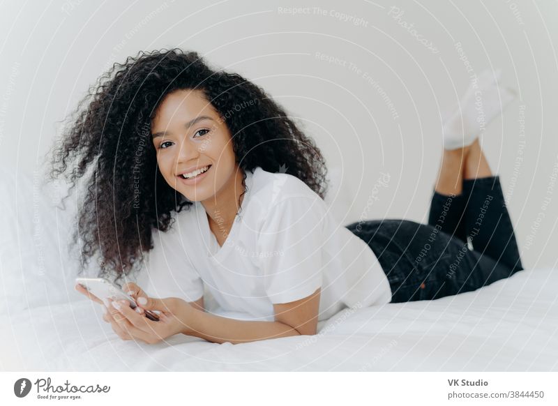 Horizontale Aufnahme einer gelockten ethnischen Frau mit angenehmem Lächeln, liegt auf einem bequemen Bett, hebt die Beine an, benutzt ein modernes Mobiltelefon, liest Textnachrichten, genießt eine Hochgeschwindigkeits-Internetverbindung. Menschen und Schlafenszeit