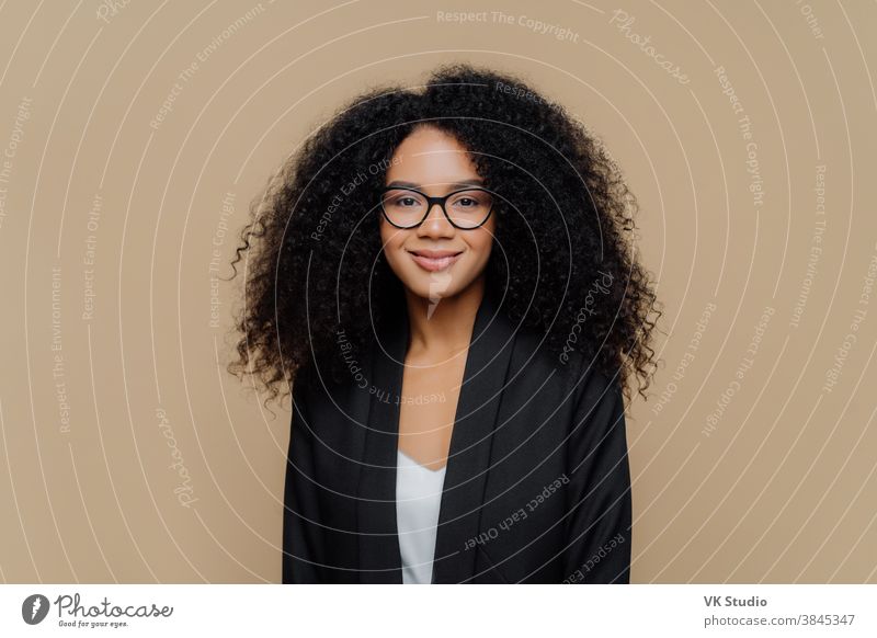 Porträt einer schönen Afroamerikanerin mit knackigem Haar, gekleidet in eleganter schwarzer Jacke, transparente Brille, schaut direkt in die Kamera mit sanftem Lächeln, trägt eine optische Brille, die isoliert an einer braunen Wand angebracht ist