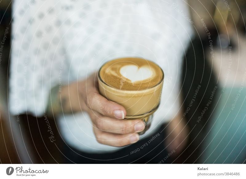 Glas Milchkaffee mit Latte Art Herz in der Hand einer Frau mit weißer Bluse Latte Macchiato milchschaum Barista Espresso Frühstück Kaffee Café café au lait