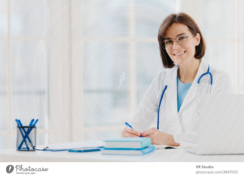 Berufstätige Ärztin schreibt Notizen, posiert mit Laptop am Schreibtisch im Büro, trägt weißen Kittel, Brille und Phonendoskop um den Hals, blickt durch medizinische Dokumente. Konzept der Gesundheitsversorgung