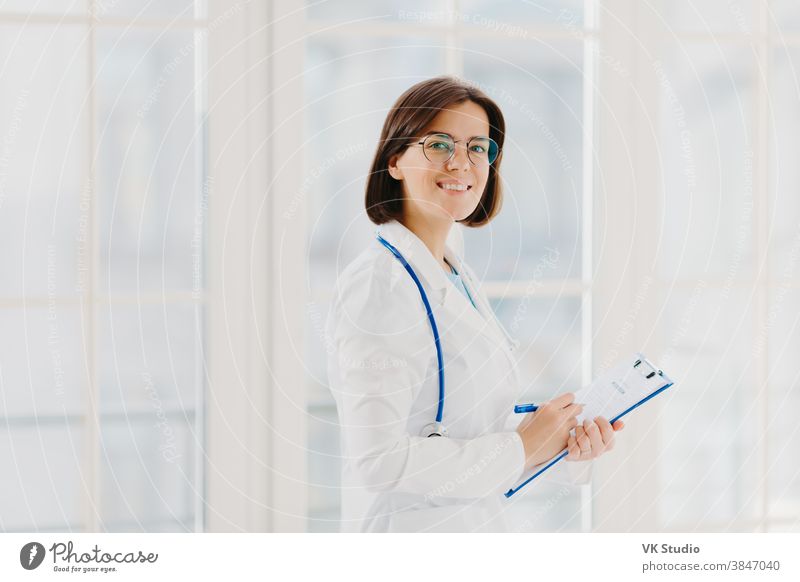 Horizontale Aufnahme der Ärztin füllt medizinisches Formular am Klemmbrett aus, steht im Haus, trägt eine runde Brille, einen weißen Kittel und ein Stethoskop. Allgemeinmedizinerin schreibt Notizen, konsultiert Patienten