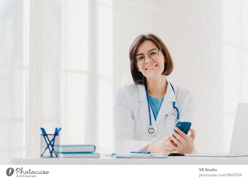 Horizontale Ansicht eines lächelnden professionellen Arztes arbeitet in der Klinik, posiert in einem modernen Krankenhausbüro mit elektronischen Geräten, verschickt Textnachrichten auf dem Handy während der Arbeit. Gesundheitsversorgung, Technologiekonzept