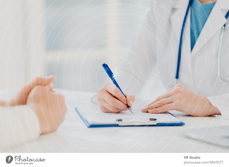 Unbekannte Ärztin schreibt Rezept auf Papiere in der Zwischenablage, konsultiert Patientin zur Heilung von Krankheit, posiert am Schreibtisch vor unscharfem weißem Hintergrund und füllt medizinisches Formular aus. Diagnose der Symptome