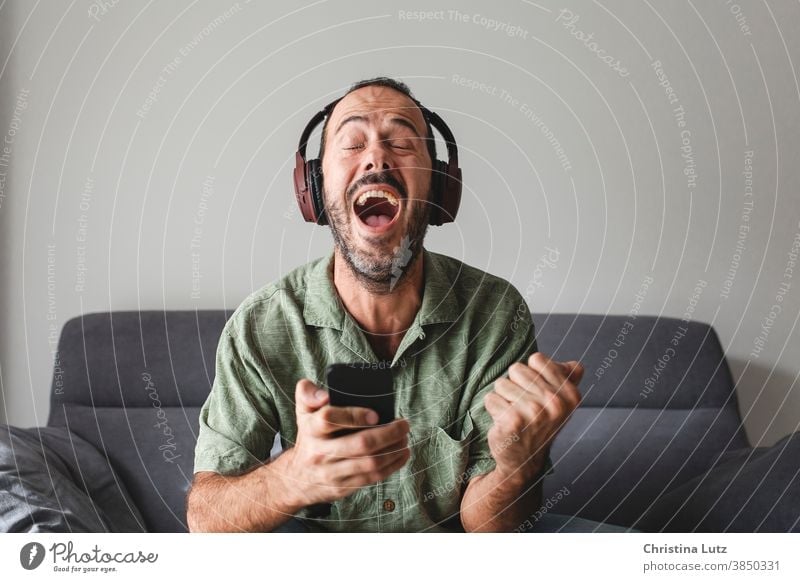 Mann, der mit Handy und Kopfhörer auf dem Sofa sitzt und vor Freude schreit, während er Sportwetten gewinnt vor Freude schreien Smartphone Ergebnisse