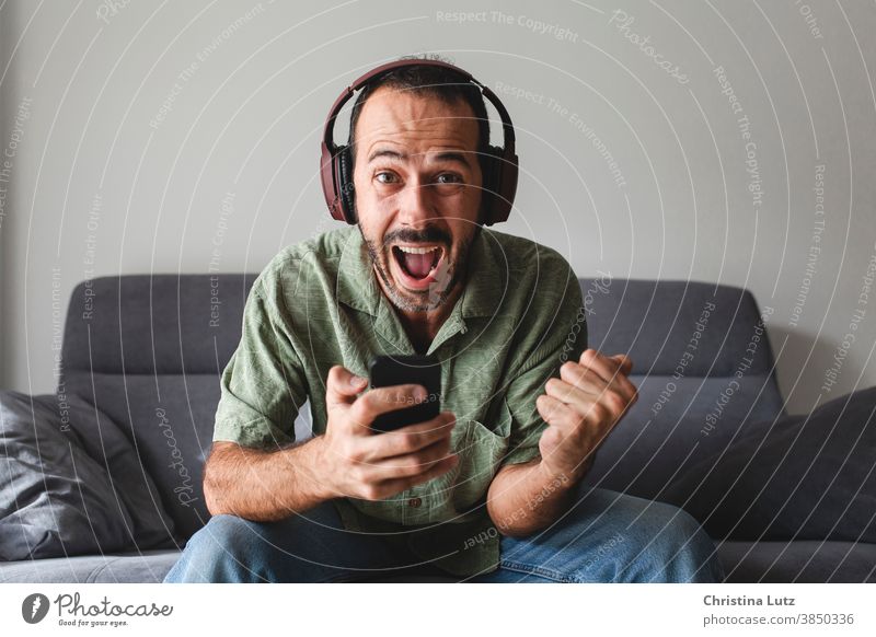 Mann, der auf dem Sofa sitzt und vor Freude schreit, während er mit dem Handy Sportwetten gewinnt vor Freude schreien Telefon geballte Faust Schreien Kopfhörer