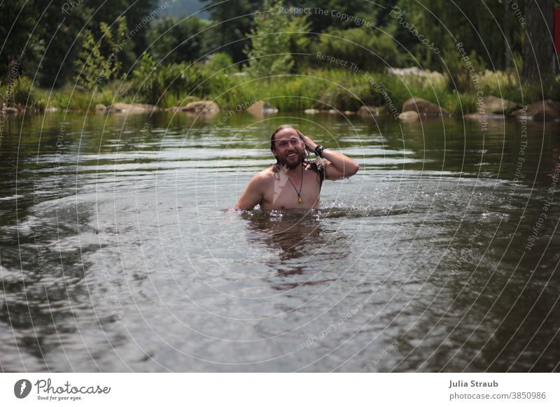 Langhaariger Mann badet in einem Natursee im Sommer Urlaub Badesee See Wasser lange Haare Vollbart Kette Armband Lächeln natürlich Seeufer Steine Bäume Waldrand