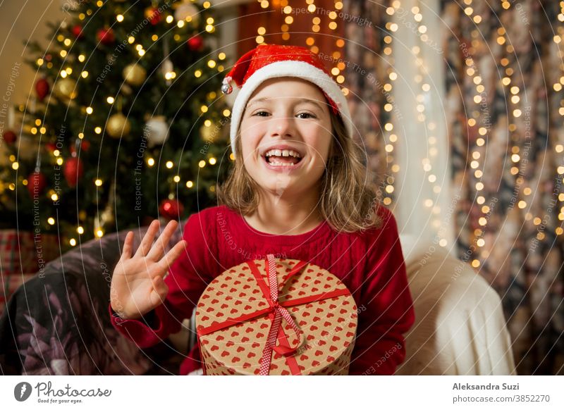 Ein glückliches Kind mit roter Nikolausmütze feiert Weihnachten. Ein süßes Mädchen hält ein verpacktes Geschenk in der Hand, lächelt und winkt mit der Hand, sitzt in einem mit Weihnachtslichtern geschmückten Baumzimmer. Fröhliche Feiertage