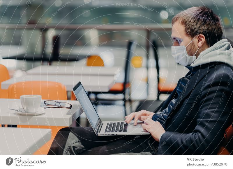 Innenaufnahme eines männlichen Freiberuflers arbeitet freiberuflich an modernem Laptop-Computer, trägt medizinische Maske gegen Coronavirus, posiert im Café, konzentriert am Bildschirm, liest Nachrichten, angeschlossen an drahtloses Internet