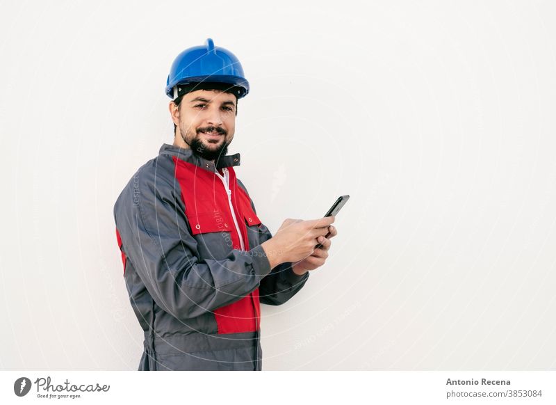 Bärtiger Arbeiter Mann mit Smartphone schaut lächelnd in die Kamera Uniform Schutzhelm Pandemie Virus weißer Hintergrund Wand Kinnbart Vollbart arabisch 40s