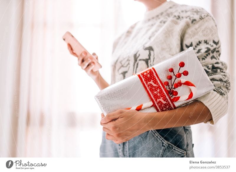 Junge unerkennbare Frau, die ihr Mobiltelefon in einer Geschenkbox aus Metallpapier mit rotem Zwirn und Süßigkeiten in der Hand hält. Weihnachts-Silvester-Geschenk.