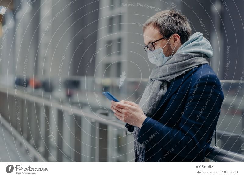 Seitliche Aufnahme eines männlichen Bloggers, der Textnachrichten verschickt und Kommentare von Anhängern auf modernem Mobiltelefon liest, trägt eine sterile medizinische Maske, um sich vor dem Coronavirus oder einer anderen Infektionskrankheit zu schützen