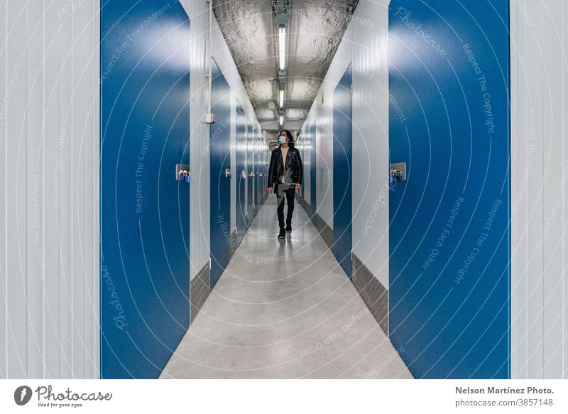 Ein spanischer Mann in Lederjacke geht durch einen Flur mit blauen Türen und weißen Wänden. Mundschutz COVID19 Tiefe Lagerraum Lifestyle cool jung Arbeit