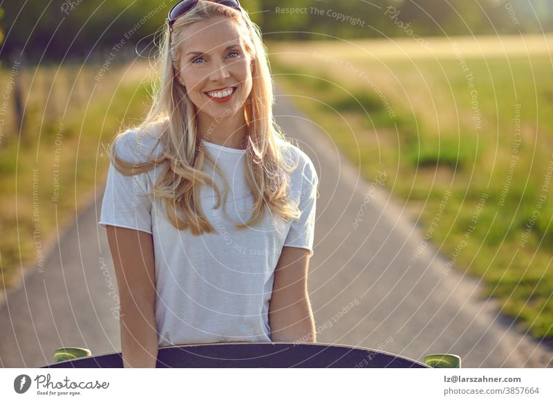 Porträt einer fitten schönen Frau mittleren Alters mit aktivem Lebensstil, die lächelt und in die Kamera schaut, während sie im Sommer auf einer sonnigen Straße im Park ein Longboard hält