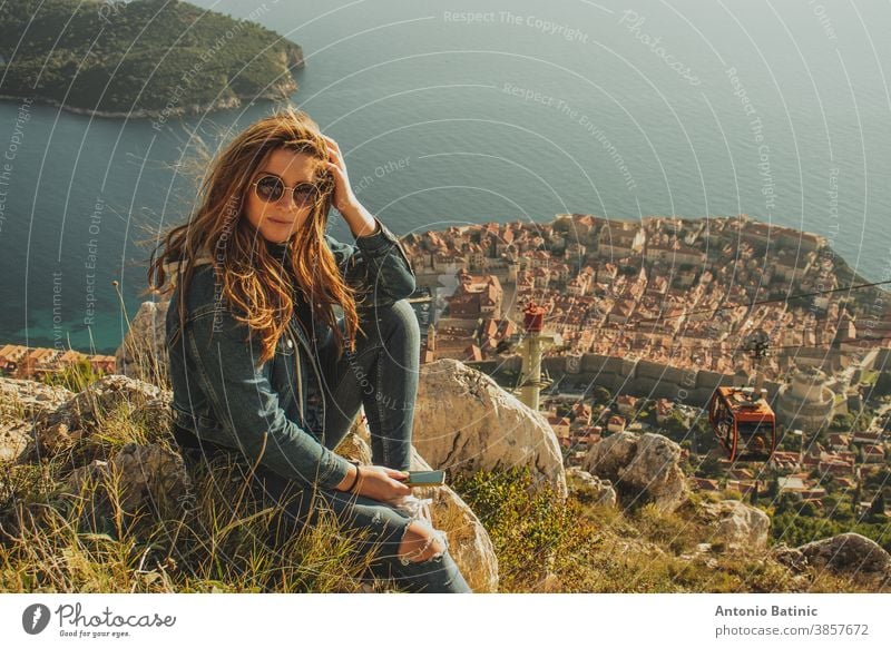 Attraktive Brünette, die auf dem Hügel Srd über der Stadt Dubrovnik sitzt und die Stadt in der Ferne beobachtet. Wind weht ihr Haar über dem alten berühmten Wahrzeichen der Stadt