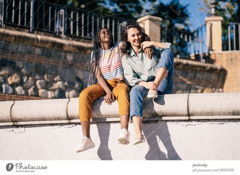 Zwei Freunde, die sich gemeinsam auf der Straße amüsieren und auf einer Stadtmauer sitzen. Frauen multiethnisch schwarz Afro-Look Mädchen Schüler zwei Spaß