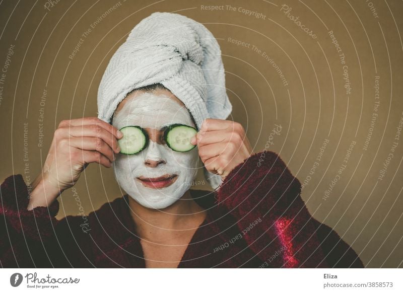 Frau mit Bademantel und Handtuch auf dem Kopf genießt entspannt eine Gesichtsmaske und hält sich zwei Gurkenscheiben vor die Augen. Wellness, Gesichtspflege.