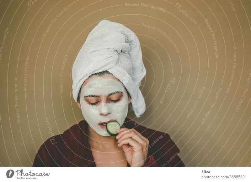 Frau mit Bademantel und Handtuch auf dem Kopf genießt entspannt eine Gesichtsmaske und nascht dabei eine Gurkenscheibe. Wellness, Gesichtspflege. Gurkenscheiben