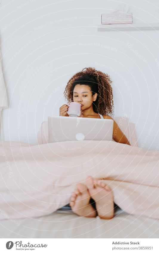 Ethnische Frau mit Tasse Kaffee und Laptop sitzt auf dem Bett Schlafzimmer trinken gelangweilt zu Hause sich[Akk] entspannen ruhen Freizeit jung Afroamerikaner