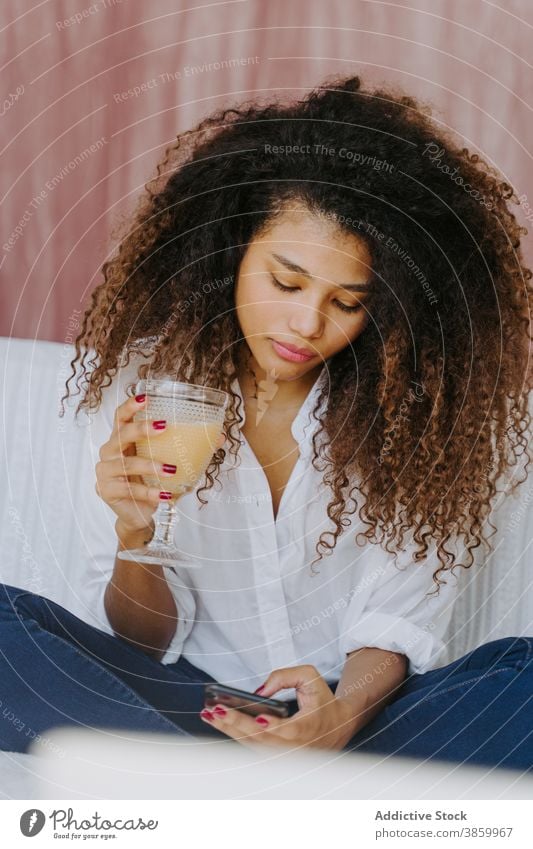 Ethnische Frau trinkt Saft und surft auf dem Smartphone zu Hause trinken benutzend Browsen Mobile Kälte Telefon Apparatur jung ethnisch Afroamerikaner schwarz