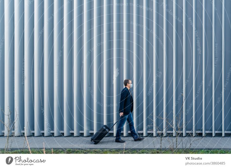 Horizontale Panoramaaufnahme eines Mannes, der im eigenen Land wegen Quarantäne und Pandemie in der Welt eintrifft, geht mit dem Koffer, stellt sich im Freien gegen einen Metallzaun, trägt Gesichtsmaske