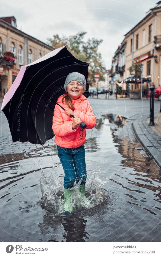 Kleines Mädchen mit großem Regenschirm in der Hand springt an einem verregneten, düsteren Herbsttag in die Pfütze im Freien wenig saisonbedingt fallen Kindheit