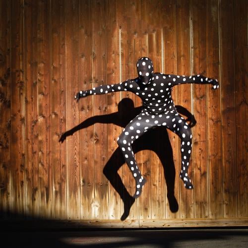 Menschliche Gestalt im gepunktetem Morphsuitkostüm , Sprung in die Luft mit Schattenwurf auf eine Holzwand Morphsuite ganzkörperanzug Punkte im Sprung androgyn
