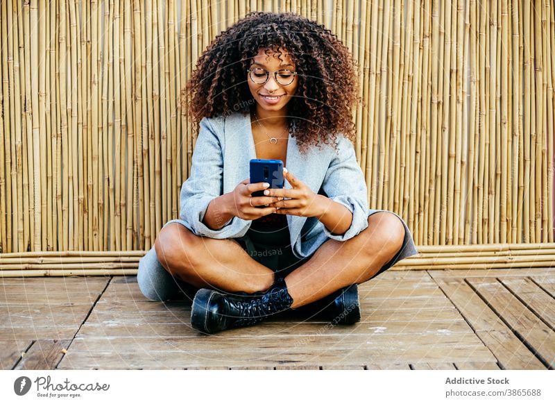 Fröhliche schwarze Frau mit Smartphone in der Nähe von Bambus Wand benutzend ruhen Stil Stock hölzern Lächeln Beine gekreuzt jung Outfit Wochenende trendy