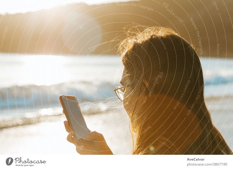 Junge Frau überprüft sein Handy am Strand während eines Sonnenuntergangs mit Kopierraum auf bunten Tönen Person Lächeln Drahtlos Beteiligung