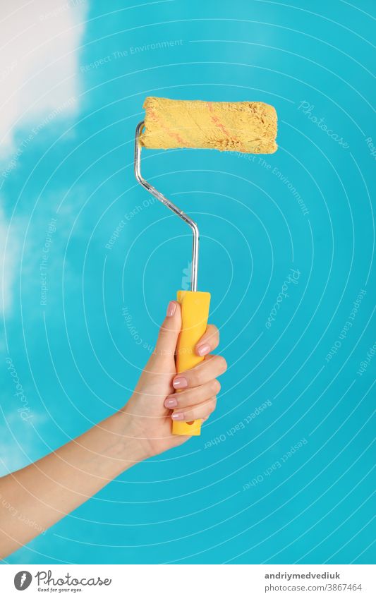 Nahaufnahme einer weiblichen Hand, die eine gelbe Farbwalze über einen blauen Hintergrund hält - Reparatur-, Konstruktions- und Bauwerkzeugkonzept. Bequemes und vielseitiges Werkzeug zum Bemalen von Wänden.