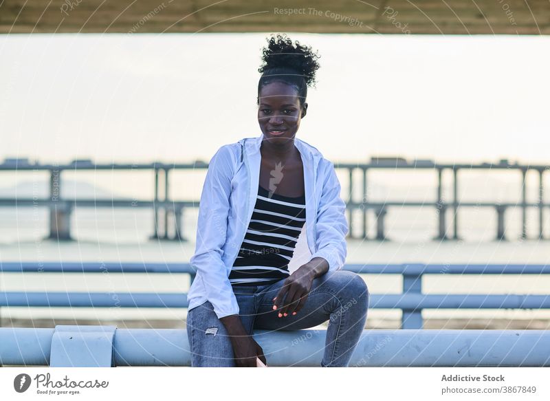 Glückliche ethnische Frau ruht auf Zaun in der Nähe von Meer Stauanlage Kälte ruhen lässig Lächeln Strandpromenade Reling jung Afroamerikaner schwarz urban