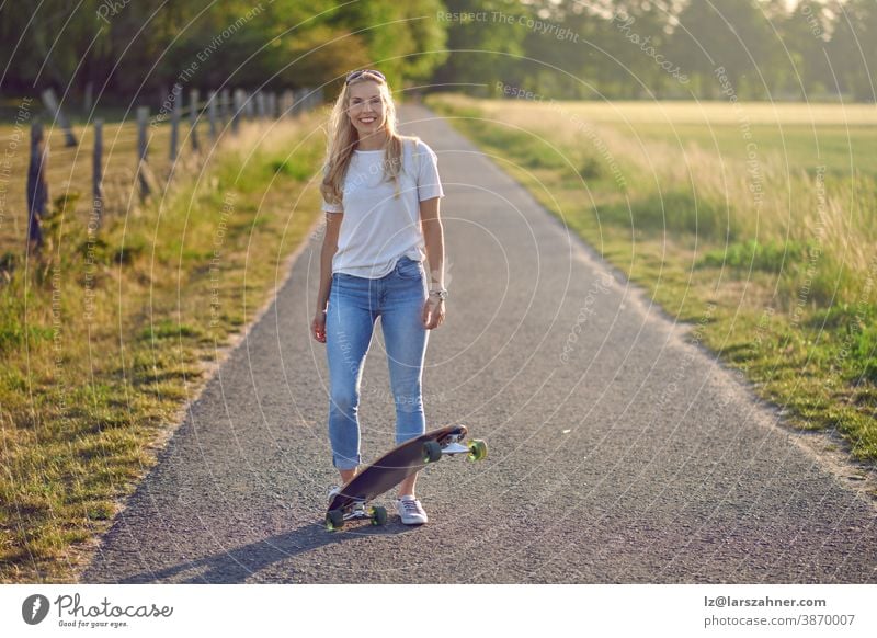 Attraktive sportliche blonde Frau mit ihrem Skateboard im Gegenlicht der Abendsonne auf einer Landstraße zwischen Feldern, die in die Kamera lächelt