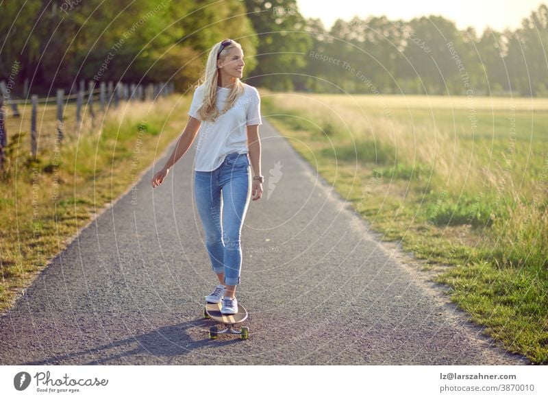 Passgenaue aktive Frau mittleren Alters spielt auf ihrem Skateboard und nähert sich der Kamera entlang einer schmalen Landstraße mit einem glücklichen Lächeln im Gegenlicht der Abendsonne